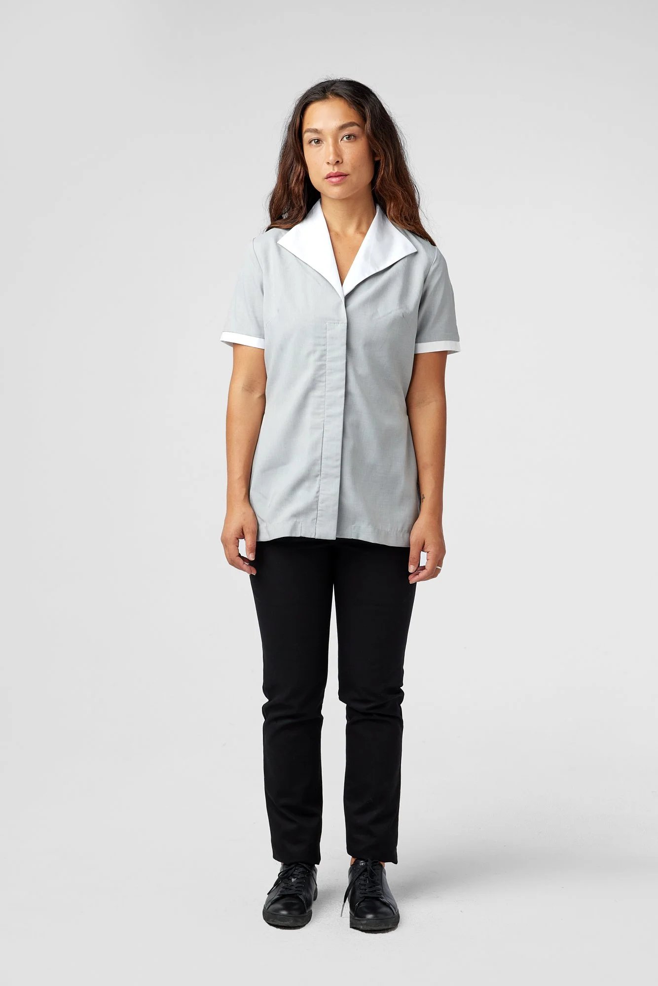 A woman with long brown hair stands facing forward, wearing a light gray short-sleeve uniform top with a white collar, black pants, and black shoes, against a plain light gray background.