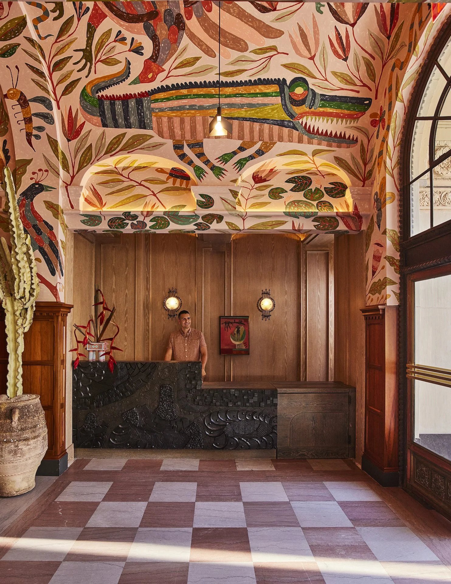 Colorful, tropical-themed hotel lobby with a mural of an alligator and foliage on the ceiling and walls. A person stands behind a dark reception desk; the space has tiled floors, wood paneling, and a large potted cactus.