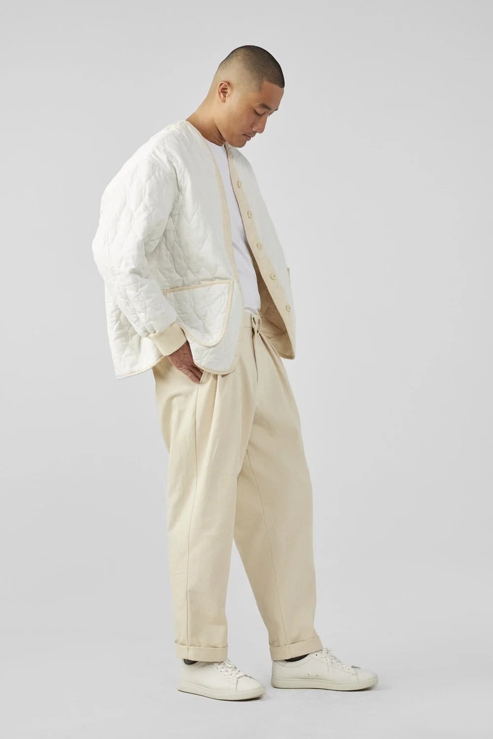 A man stands sideways wearing a white quilted jacket, a white T-shirt, loose beige pants, and white sneakers against a plain light gray background.