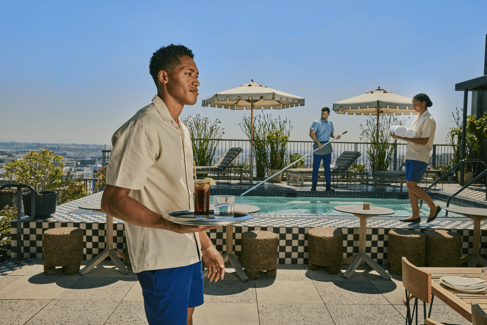 A young man carries drinks on a tray by a pool, while another person cleans the pool and a third person sets out towels under large umbrellas. Lounge chairs and plants surround the pool area on a sunny day.