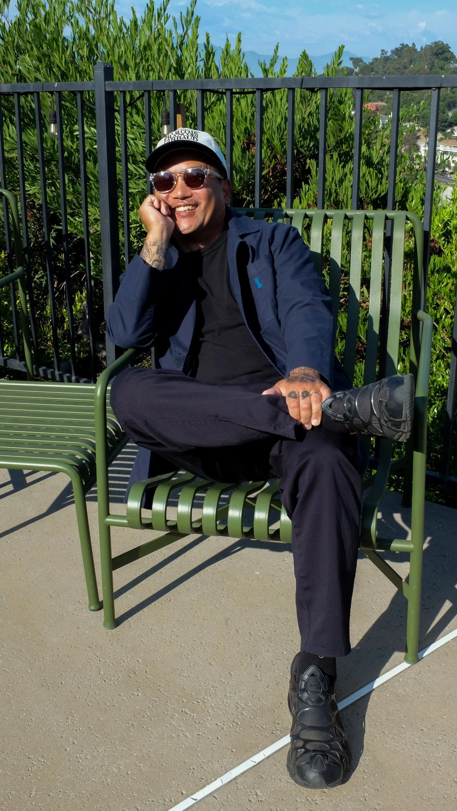 A man sitting on a green park bench outdoors, wearing sunglasses and a baseball cap, smiling, with lush plants and blue sky in the background.
