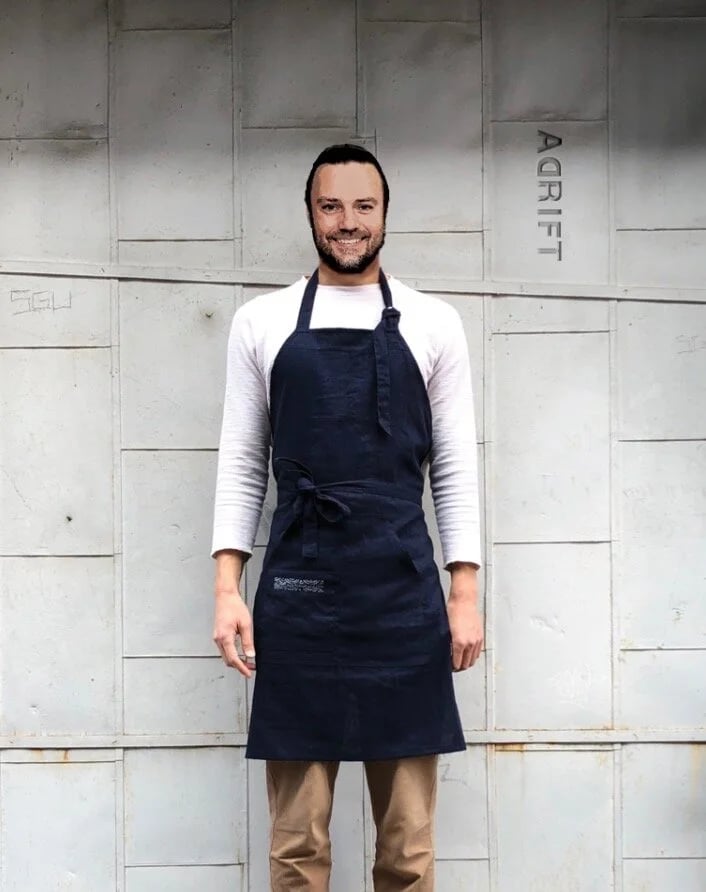 Man smiling, wearing a white long sleeve shirt and navy apron, standing in front of a concrete wall with the word "ART" on it.