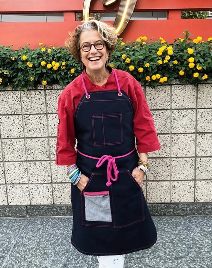 Smiling woman with curly gray hair and glasses wearing a red shirt and a dark apron with pink accents, standing in front of a brick wall with yellow flowers and a neon sign.