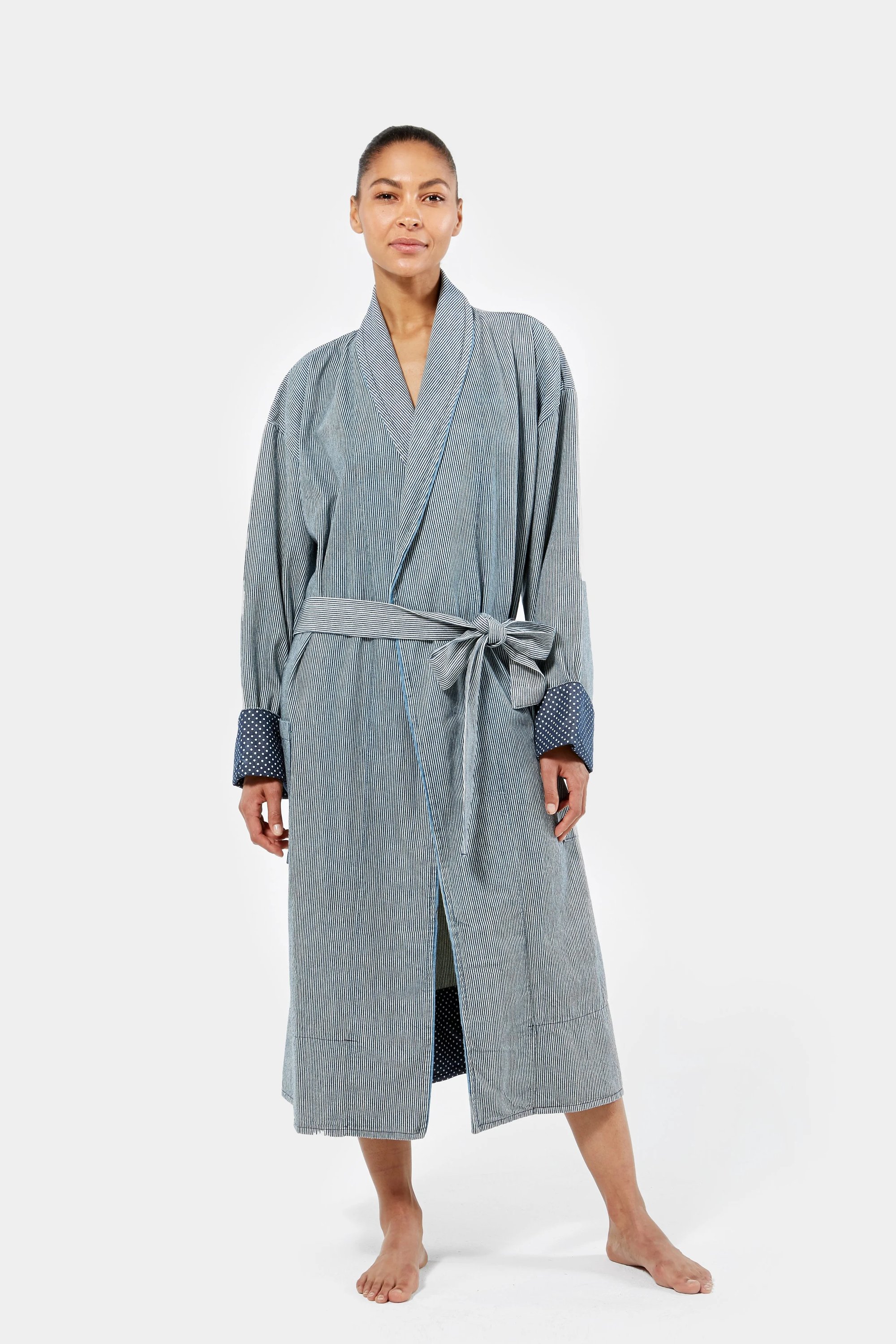 A woman standing barefoot on a white background, wearing a long, grey robe with a belt and dark cuffs.
