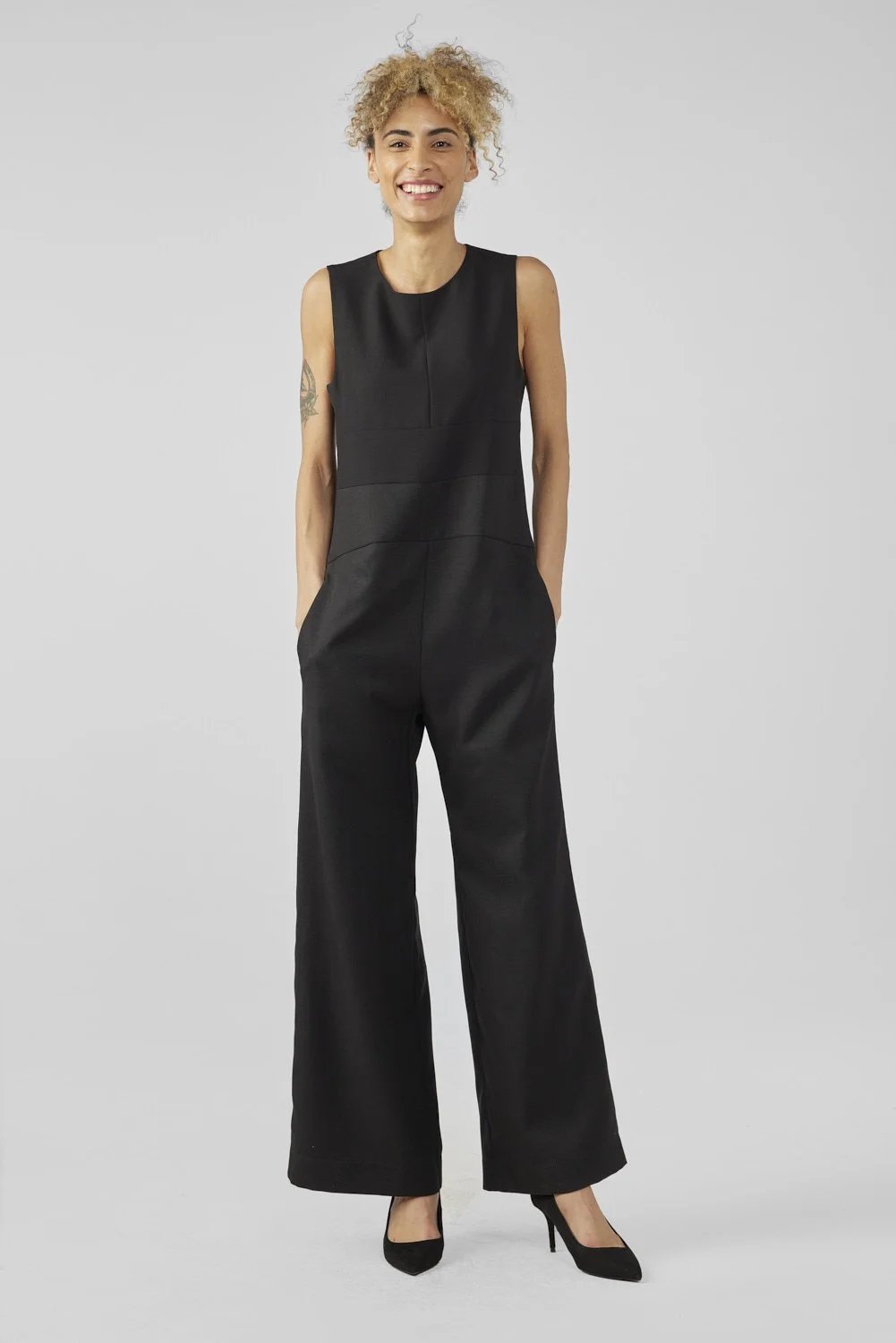 A person with curly hair wearing a sleeveless black jumpsuit and black heels stands against a plain gray background, smiling with hands in their pockets.