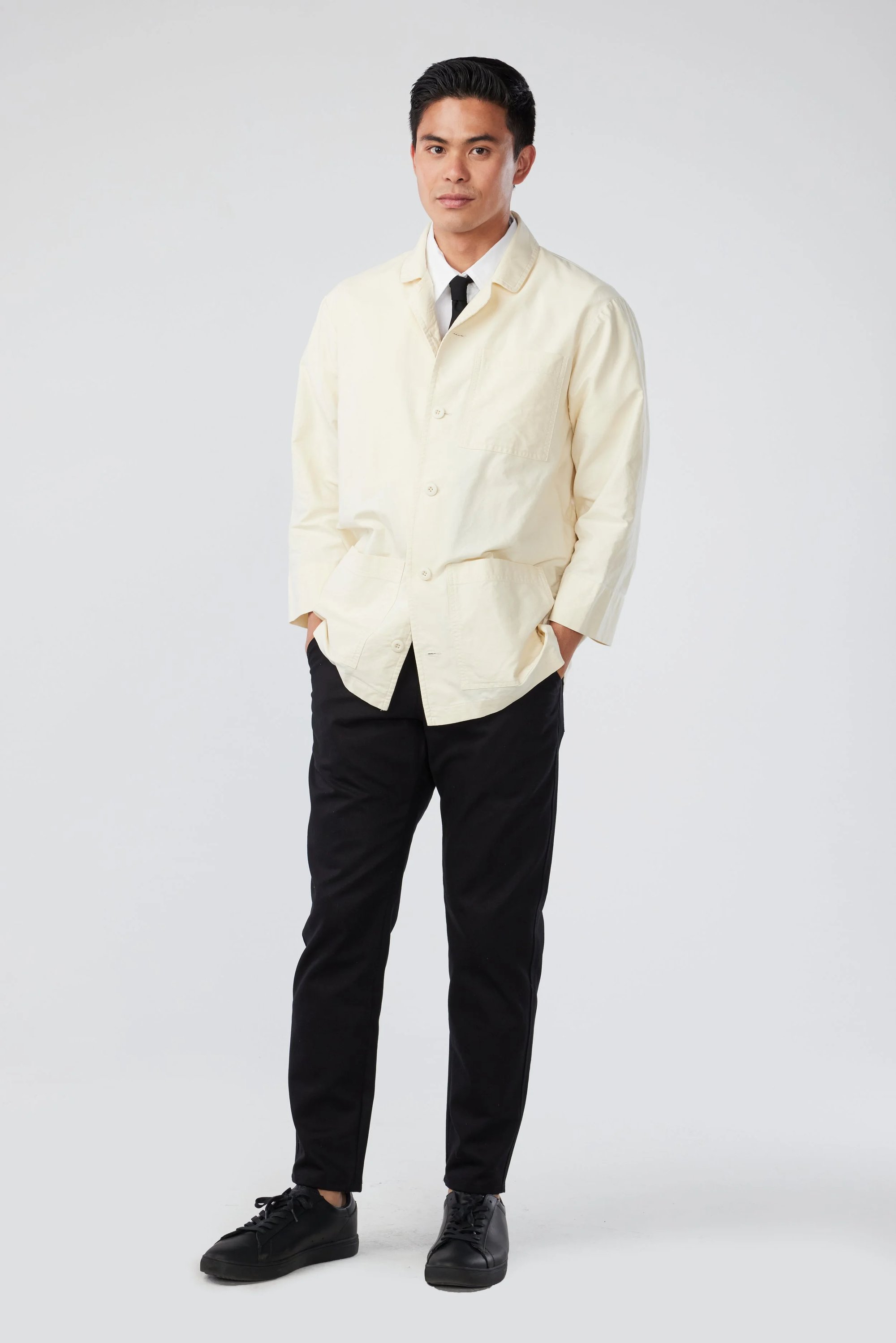 A man stands against a plain background wearing a cream-colored button-up jacket over a white shirt and black tie, paired with black pants and black shoes. His hands are in his pockets and he looks forward.