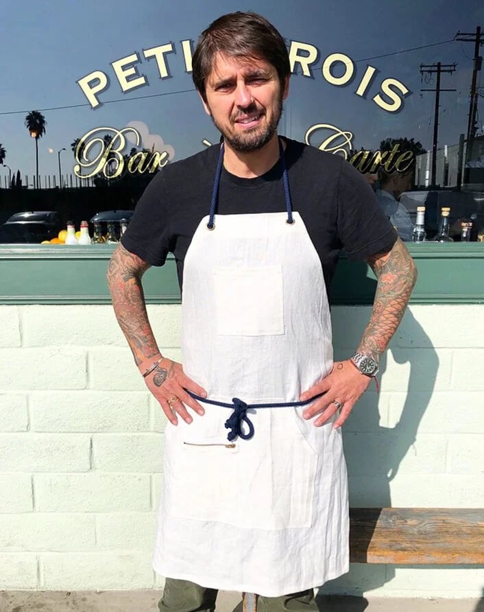 Man wearing a black t-shirt and white apron standing outside a restaurant with a sign reading "Petite Rois Bar" in the background.