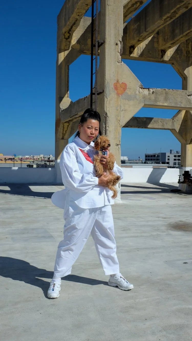 Woman in white athletic clothing holding a small brown dog on a sunny rooftop with a blue sky and urban buildings in the background.