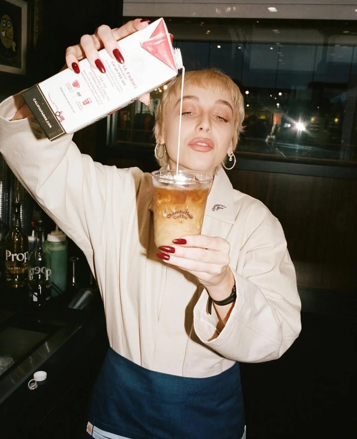 A person with short blond hair pours milk from a carton into a cup of iced coffee, standing behind a counter and wearing a light jacket and blue apron uniform.