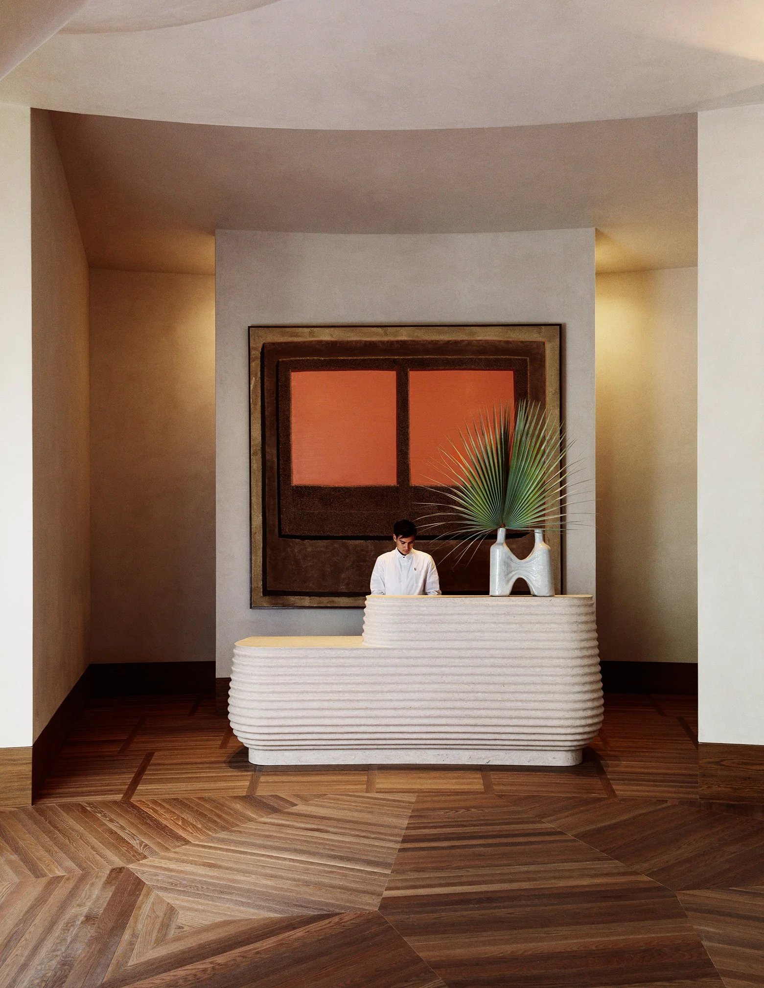 A person stands at a modern, curved reception desk with a large plant and vases on top, set against a geometric wooden floor and a wall-mounted abstract artwork with brown and orange rectangles.