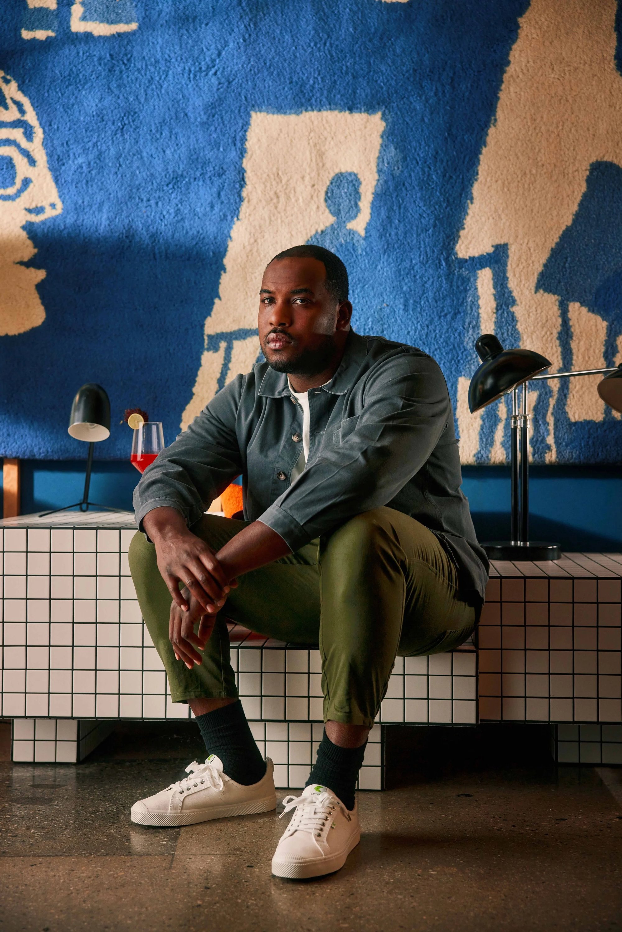 A man in a gray jacket, green pants, and white sneakers sits on a tiled bench in a modern room with a blue and beige abstract wall hanging and desk lamps behind him.