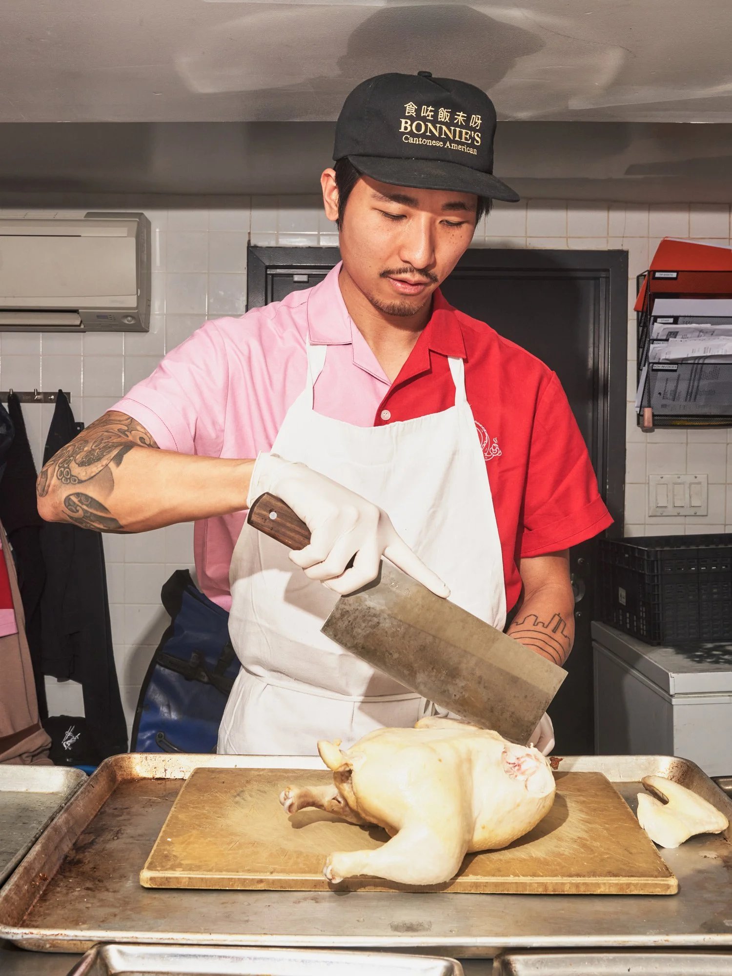 A chef in a red/pink shirt, white apron, and black cap uses a large cleaver to cut a raw chicken on a tray in a commercial kitchen.
