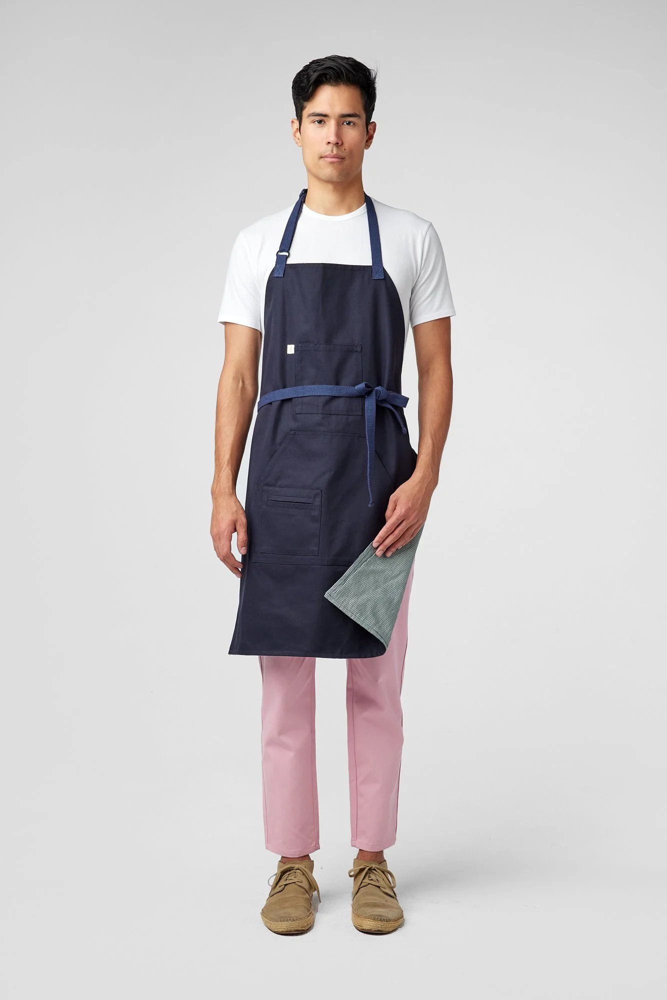 A person wearing a white t-shirt, navy blue apron, pink pants, and brown shoes stands against a plain light gray background. They are folding the bottom-left corner of the apron to reveal a gray inside lining.