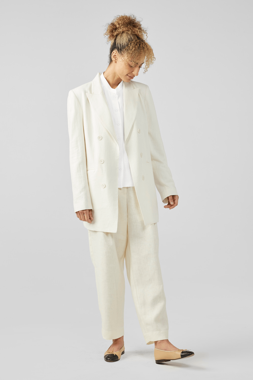 A person with curly hair wears a white double-breasted blazer, white shirt, and loose white trousers. They are looking down and smiling, with their hands relaxed by their sides. They wear beige flats with black toe caps.