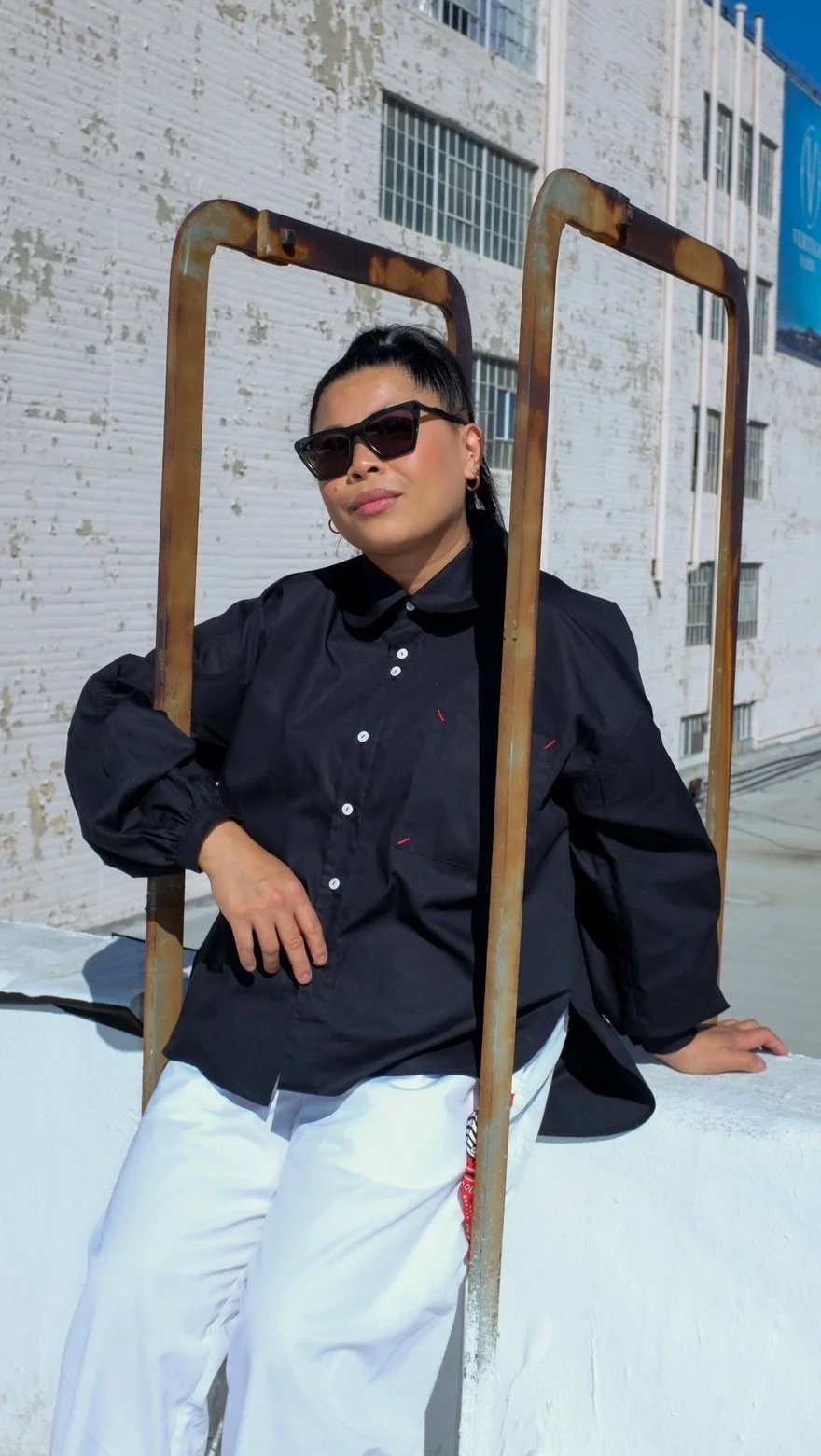 A woman wearing black sunglasses, a black long-sleeved shirt, and white pants poses outdoors against a white textured wall, with a rusty metal frame around her, under bright sunlight.