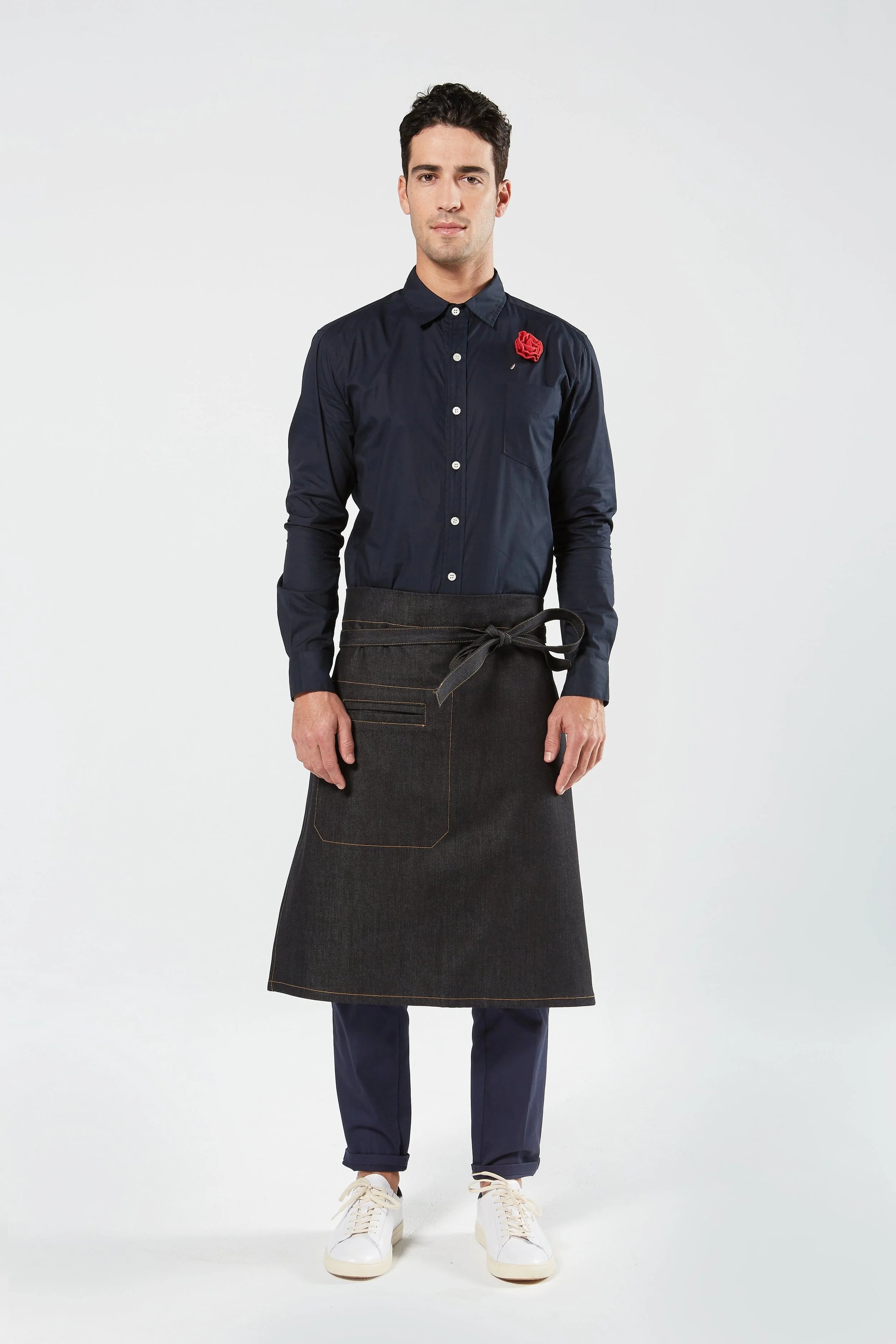 A man stands facing forward, wearing a dark button-up shirt with a red flower on the chest, a black waist apron, dark pants, and white sneakers against a plain light background.