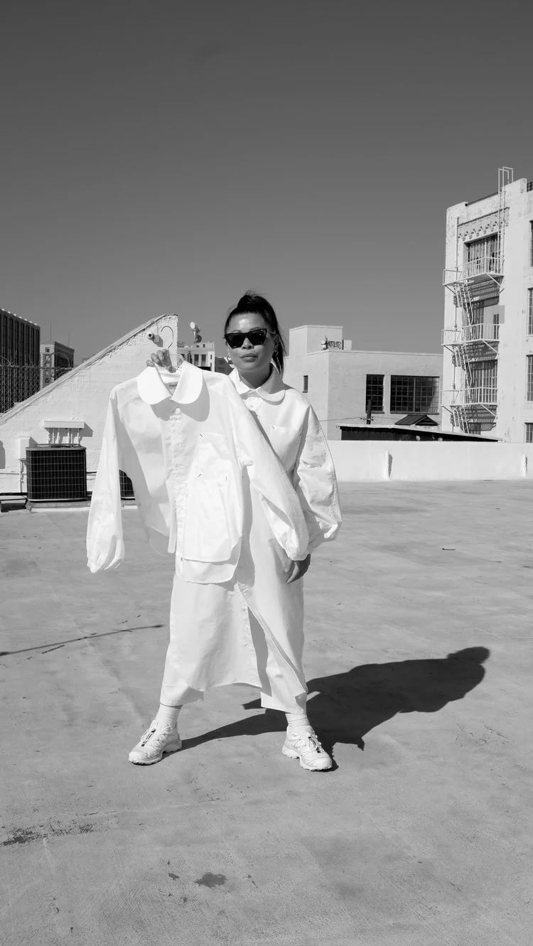 Woman wearing sunglasses holding a white jacket on a hanger on a rooftop with buildings in the background.