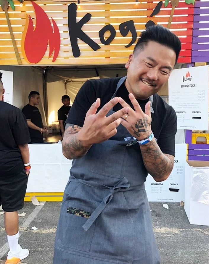 Man smiling and making a hand gesture at an outdoor food event, wearing a gray apron with a tattooed arm, in front of a colorful sign that says 'Kogi' with a flame logo.
